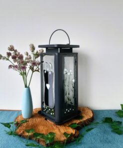 Decorative lantern with heart design on a wooden base, next to a blue vase with pink flowers, against a plain background.