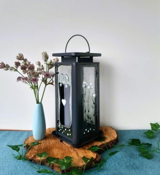 Decorative lantern with heart design on a wooden base, next to a blue vase with pink flowers, against a plain background.