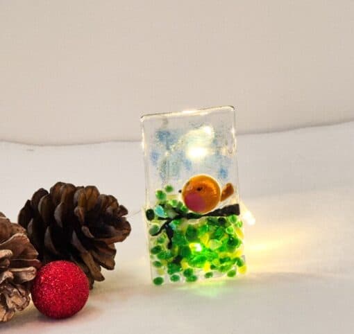 Glass ornament with a small bird and green beads, lit from inside. Two pinecones and a red ornament sit beside it.