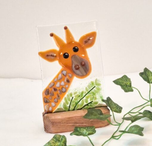 Glass artwork of a giraffe with green leaves, mounted on a wooden base, against a neutral background.