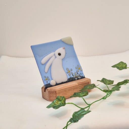 Glass art featuring a white rabbit with flowers on a blue background in a wooden stand, decorated with a green vine.