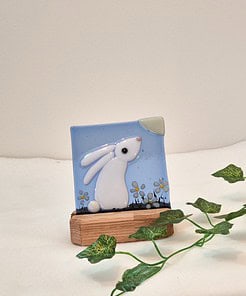 Glass artwork of a white rabbit with flowers on a blue background, set on a wooden stand, with green ivy nearby.