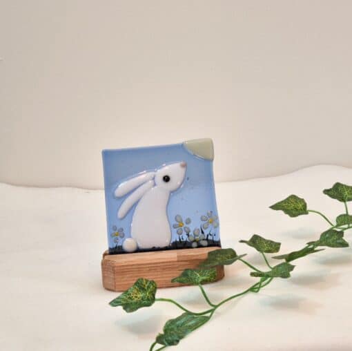 Glass artwork of a white rabbit with flowers on a blue background, set on a wooden stand, with green ivy nearby.