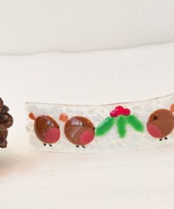 Glass ornament with red and green details next to two pine cones and a glittery red ball on a white surface.