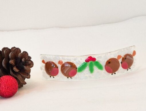 Glass ornament with red and green details next to two pine cones and a glittery red ball on a white surface.