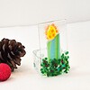 Glass candle ornament with green base and yellow flame beside pinecones and a small red glitter ball on a white background.