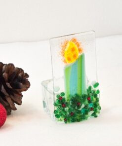 Glass candle ornament with green base and yellow flame beside pinecones and a small red glitter ball on a white background.