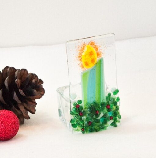 Glass candle ornament with green base and yellow flame beside pinecones and a small red glitter ball on a white background.