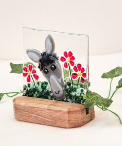 Ceramic donkey face decor with red flowers in a wooden base, surrounded by green leaves on a white background.