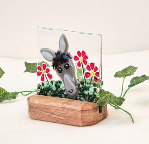 Ceramic donkey face decor with red flowers in a wooden base, surrounded by green leaves on a white background.