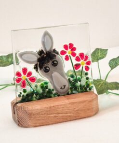 Glass art piece with a donkey face and red flowers on a wooden base, surrounded by small green leaves.