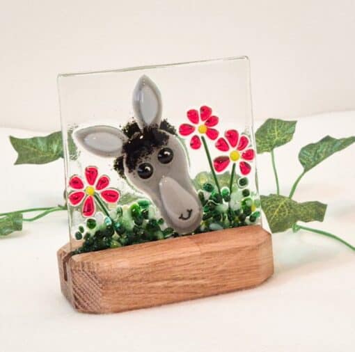 Glass art piece with a donkey face and red flowers on a wooden base, surrounded by small green leaves.