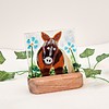 Glass horse figurine with blue flowers on a wooden stand, surrounded by green leaves against a plain background.