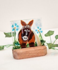 Glass horse figurine with blue flowers on a wooden stand, surrounded by green leaves against a plain background.