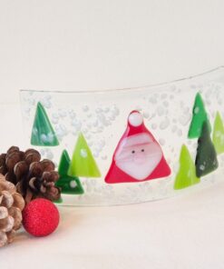 Curved glass art with a Santa figure and green trees, surrounded by pinecones and a red ornament on a white surface.