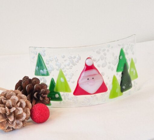 Curved glass art with a Santa figure and green trees, surrounded by pinecones and a red ornament on a white surface.