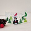 Glass art with a penguin in a Santa hat, surrounded by green trees and snow, next to pinecones and a red ornament.