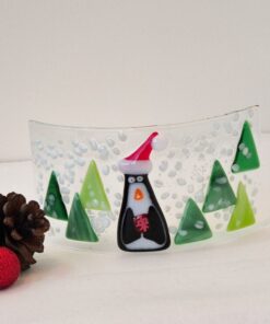 Glass art with a penguin in a Santa hat, surrounded by green trees and snow, next to pinecones and a red ornament.