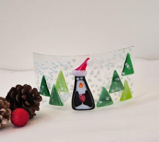 Glass art with a penguin in a Santa hat, surrounded by green trees and snow, next to pinecones and a red ornament.