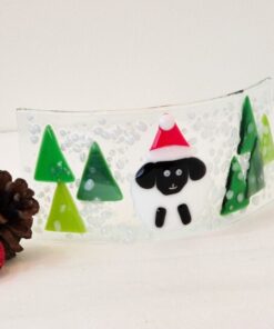 Decorative glass panel with a Santa hat-wearing sheep and green trees, placed beside pinecones and a red ornament.