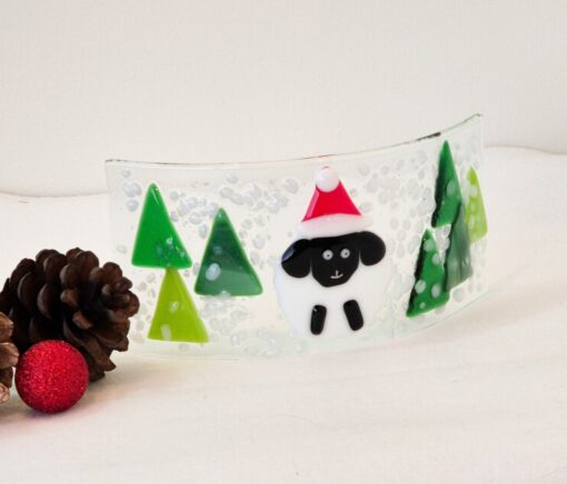 Decorative glass panel with a Santa hat-wearing sheep and green trees, placed beside pinecones and a red ornament.