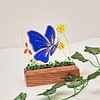 Blue butterfly stained glass art in wooden base, with green vine decoration on white background.