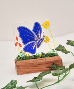 Blue butterfly stained glass art in wooden base, with green vine decoration on white background.