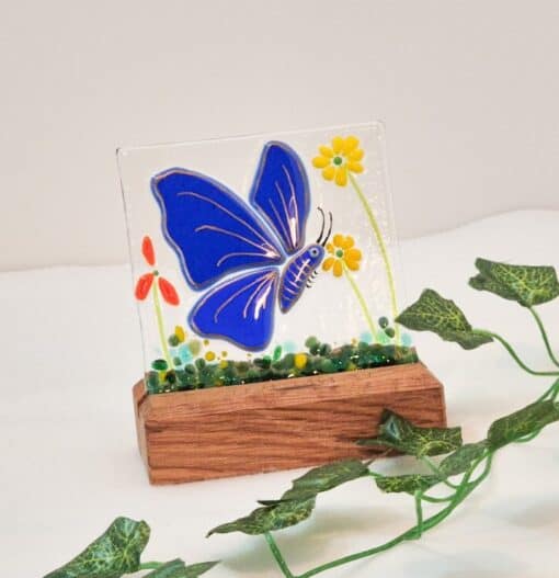 Blue butterfly stained glass art in wooden base, with green vine decoration on white background.