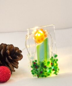 Festive glass candle ornament glows, surrounded by pine cones and a small red glittered ball on a white surface.