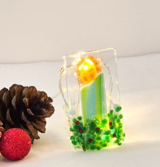 Festive glass candle ornament glows, surrounded by pine cones and a small red glittered ball on a white surface.