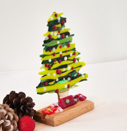 Glass Christmas tree with colorful ornaments on a wooden base, surrounded by pinecones and a red glittery ball.