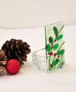 Glass votive holder with holly design beside pinecones and a red glittery ornament on a white background.