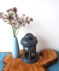 A blue vase with purple flowers and a grey lantern sit on a wooden slab atop a blue surface.