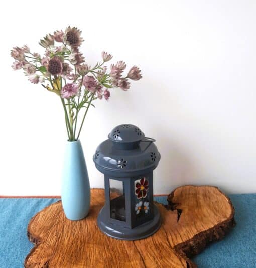 A blue vase with purple flowers and a grey lantern sit on a wooden slab atop a blue surface.