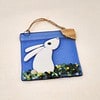 Glass ornament of a white rabbit on blue background with a crescent moon and greenery, hanging by a rope loop.