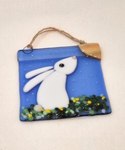 Glass ornament of a white rabbit on blue background with a crescent moon and greenery, hanging by a rope loop.