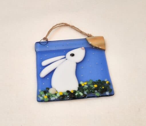 Glass ornament of a white rabbit on blue background with a crescent moon and greenery, hanging by a rope loop.