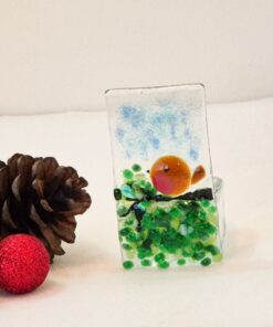 Glass ornament with a robin design next to pinecones and a red glitter ball on a white surface.