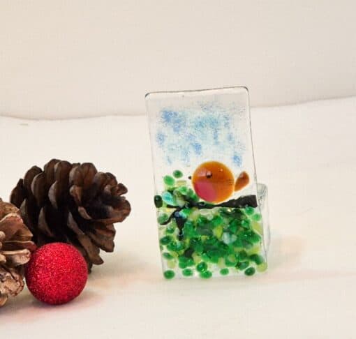 Glass ornament with a robin design next to pinecones and a red glitter ball on a white surface.