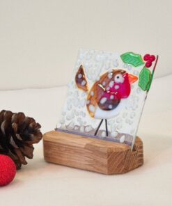 Glass artwork of a robin with a red breast and holly on a wooden stand, next to pine cones and a red ornament.