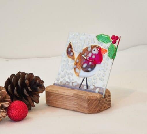 Glass artwork of a robin with a red breast and holly on a wooden stand, next to pine cones and a red ornament.