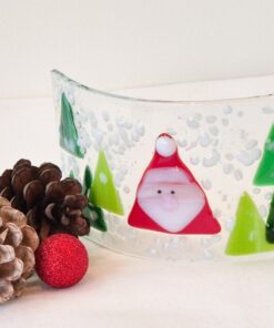 Curved glass panel with Santa and trees, surrounded by pine cones and a red ornament on a white surface.