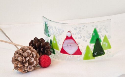 Curved glass panel with Santa and trees, surrounded by pine cones and a red ornament on a white surface.