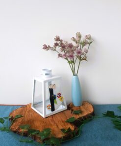 White lantern with decorative figures and flowers, next to lavender flowers in a blue vase on a wooden slab, with green vines.