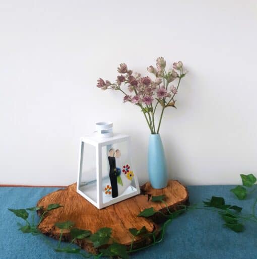 White lantern with decorative figures and flowers, next to lavender flowers in a blue vase on a wooden slab, with green vines.