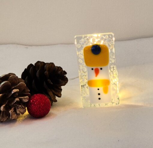 Glass snowman ornament with string lights, pine cones, and a red glitter ball on a white surface.