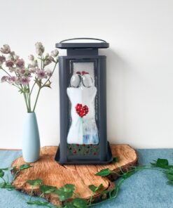 Decorative lantern with red berry design on a wooden base, beside a light blue vase with purple flowers and green vines.