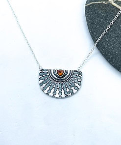 Silver half-circle pendant necklace with engraved sunburst design and amber gemstone on a white background with a dark stone accent.