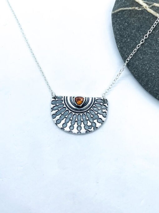 Silver half-circle pendant necklace with engraved sunburst design and amber gemstone on a white background with a dark stone accent.