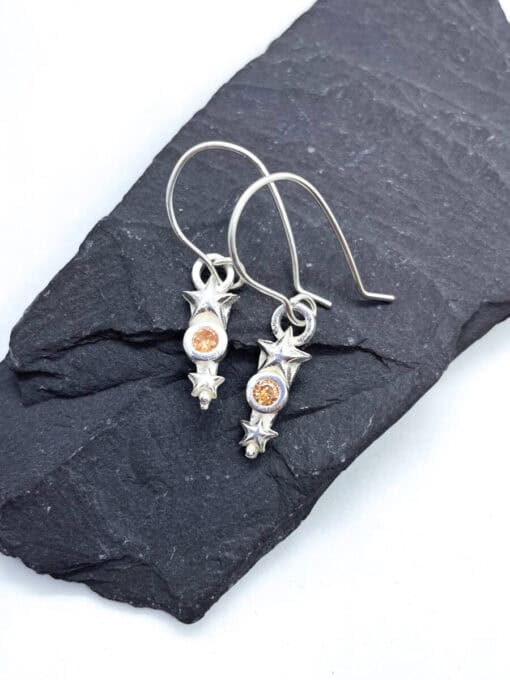 Silver star-themed earrings with orange gemstones displayed on a dark slate background.